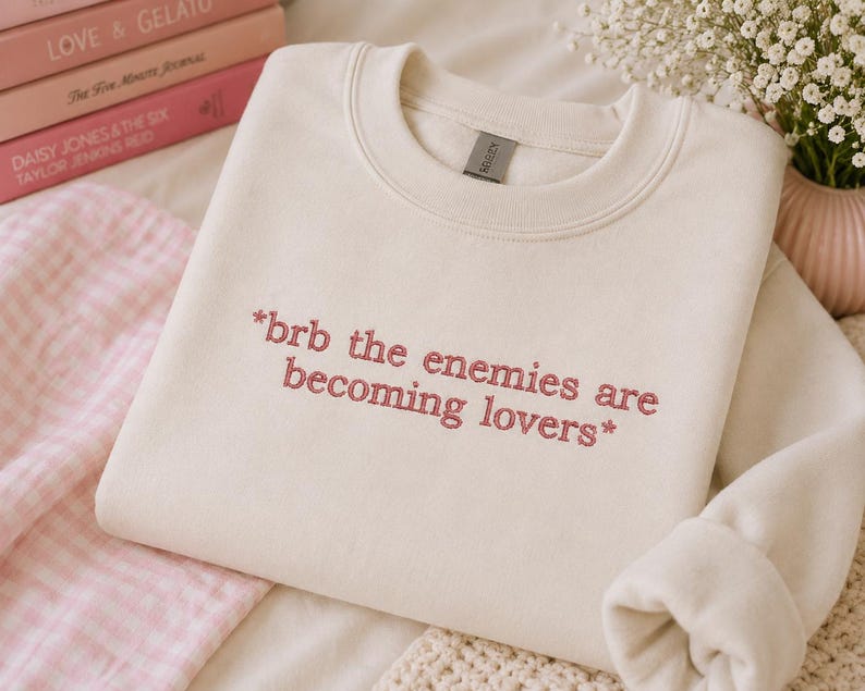 May include: Cream-colored sweatshirt with the embroidered text "*brb the enemies are becoming lovers*". The crewneck sweatshirt is folded, with the sleeves visible. A stack of pink books and a small vase with white flowers are in the background.