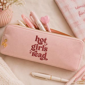 Op de afbeelding: Roze etui met de geborduurde tekst "hot girls read" in bordeauxrood. De etui is open en toont pennen en een hartvormige penpunt. Een notitieboek met de woorden "romance fantasy coffee" is zichtbaar.