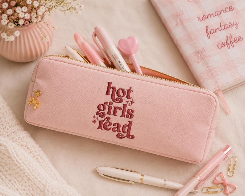 Op de afbeelding: Roze etui met de geborduurde tekst "hot girls read" in bordeauxrood. De etui is open en toont pennen en een hartvormige penpunt. Een notitieboek met de woorden "romance fantasy coffee" is zichtbaar.