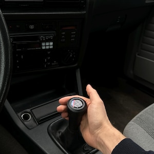 May include: Black gear shift knob with a multi-coloured emblem, held by a hand inside a car. The interior is dark with a black dashboard and air vents. The car's interior is grey and black.