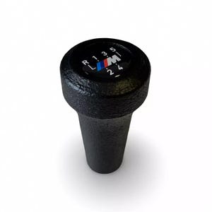 May include: Black gear stick knob with the BMW M logo. The knob has a textured surface and displays the gear shift pattern: R, 1, 3, 5, 2, 4. The BMW M logo features the colours blue, dark blue, and red.