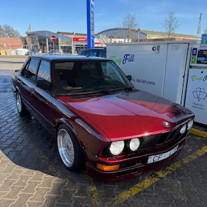 Op de afbeelding: Een bordeauxrode vintage BMW sedan geparkeerd bij een benzinestation. De auto heeft een klassiek ontwerp met chromen accenten en een zwart dak. De kentekenplaat luidt CF-476.