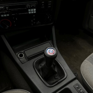 Puede incluir: Interior negro de un coche con una palanca de cambios con el logotipo de BMW. El pomo tiene un emblema circular en rojo, azul y blanco. La consola circundante también es negra.