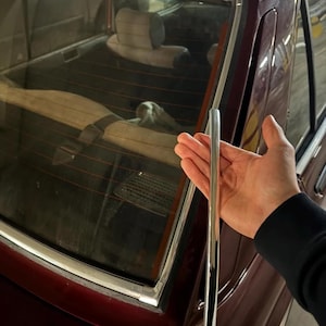 May include: A shiny, chrome trim piece held near the rear window of a maroon car. The car's interior, including the back seat and headrests, is visible through the glass. The trim appears to be a replacement part.