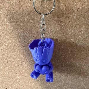 May include: A blue keychain with a 3D-printed figure of a character holding a heart. The keychain has a silver ring and chain, and is attached to a green pin on a corkboard background. The figure is approximately 5 cm tall.