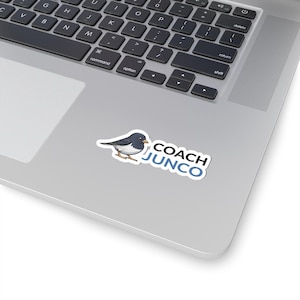 May include: A sticker featuring a cartoon bird with a blue-gray head and back, white belly, and yellow beak. The sticker also includes the text "COACH JUNCO" in a white font with a blue outline.