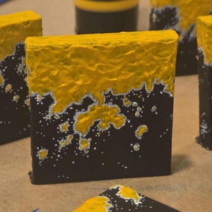 May include: Several small, square blocks with a textured yellow top and black base. The yellow paint is thick and uneven, with silver speckles and lines separating the two colours. The blocks are arranged closely together.