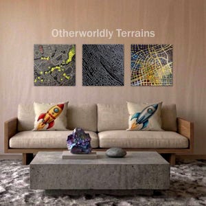 May include: Three abstract map art pieces hang above a beige sofa with rocket ship pillows. The maps feature intricate line patterns in gray, black, and gold. The text "Otherworldly Terrains" is above the art.