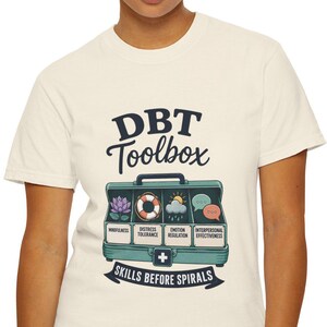 May include: A cream-colored short-sleeve t-shirt featuring a graphic of a teal toolbox labeled DBT Toolbox. The toolbox contains four compartments with icons for mindfulness, distress tolerance, emotion regulation, and interpersonal effectiveness.