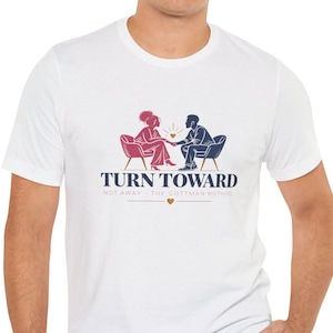 May include: A white short-sleeved t-shirt featuring a graphic of two people sitting in chairs facing each other, holding hands. The text reads TURN TOWARD NOT AWAY - THE GOTTMAN METHOD, with a small gold heart icon centered below the text.