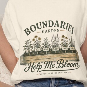 Könnte beinhalten: Ein cremefarbenes T-Shirt mit einer Grafik eines hölzernen Pflanzkastens mit verschiedenen Pflanzen. Der Text lautet "BOUNDARIES GARDEN" über den Pflanzen, "Help Me Bloom" darunter und "WATER YOUR BOUNDARIES" am unteren Rand. Jede Pflanze hat ein Schild mit der Aufschrift "NO", "REST", "SPACE", "CLARITY", "LIMITS" oder "PEACE".
