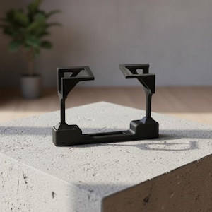 Op de afbeelding: Een zwart, geometrisch object met een rechthoekige basis en twee opstaande steunen, elk met een platte bovenkant. Het object staat op een lichtgrijze, getextureerde betonnen blok. De achtergrond omvat een raam, een plant in een pot en een glimp van een modern interieur.