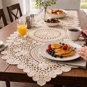 May include: A wooden dining table set for breakfast, featuring a cream-colored lace doily, plates of pancakes, fruit, and butter, along with a glass of orange juice and a cup of coffee. The table is surrounded by wooden chairs.