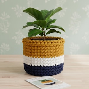 May include: A three-tone crochet plant pot in mustard yellow, white, and navy blue, holding a green plant. The pot is on a wooden surface, with a small card featuring a similar design. The pot is handmade.
