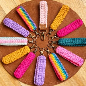 以下が含まれることがあります： 茶色の表面に円形に配置された、カラフルな手編みのリストレットキーホルダーのコレクション。キーホルダーは、ピンク、イエロー、パープル、レインボーのストライプなど、さまざまな色があり、それぞれに金属製の留め金が付いています。