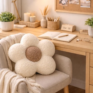 Op de afbeelding: Een bloemvormig decoratief kussen met een crèmekleurige buitenkant en een bruin midden ligt op een grijze stoel. Een crèmekleurige deken is over de stoel gedrapeerd. De scène omvat een houten bureau met een lamp, kaarsen en een prikbord.