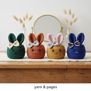 May include: Four crocheted bunny-shaped containers in green, orange, yellow, and blue. Each has upright ears and a bow. The containers are on a wooden surface with a neutral background. The text "yarn & pages" is at the bottom.