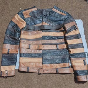 May include: A jacket constructed from horizontal panels of black, dark brown, and tan leather. The garment features a collarless neckline and multiple adjustable straps with metal buckles across the chest, waist, and sleeves for a secure fit.