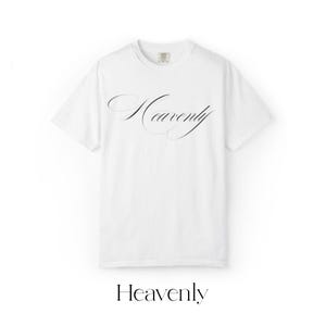 Könnte beinhalten: Weißes T-Shirt mit dem Wort "Heavenly" in eleganter schwarzer Schrift auf der Vorderseite. Das Shirt hat einen klassischen Rundhalsausschnitt und kurze Ärmel. Das Wort "Heavenly" ist auch am unteren Bildrand aufgedruckt.