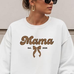 Może przedstawiać: Biała bluza z napisem "Mama" w zwierzęcym wzorze, kokardą w zwierzęcym wzorze i rokiem "2026". Bluza ma długie rękawy.