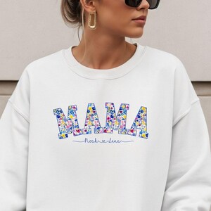 Może przedstawiać: Biała bluza z napisem "MAMA" w kolorowym kwiatowym nadruku na piersi. Poniżej napis "nosh & dear". Bluza ma swobodny, wygodny styl.