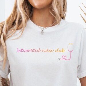 Può includere: T-shirt bianco con la scritta "Introverted nurse club" in rosa e una grafica di stetoscopio rosa e giallo. La modella indossa pantaloncini di jeans e un cardigan beige.