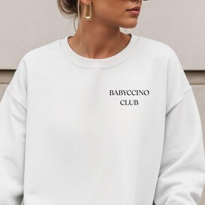 Może przedstawiać: Biała bluza z okrągłym dekoltem z czarnym napisem "BABYCCINO CLUB". Bluza jest w jednolitym białym kolorze i ma długie rękawy. Zdjęcie przedstawia bluzę na osobie.