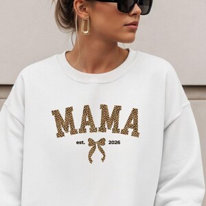 Może przedstawiać: Biała bluza z napisem "MAMA" w panterkę na piersi. Pod napisem "MAMA" znajduje się napis "est. 2026" i grafika kokardy. Bluza ma długie rękawy.