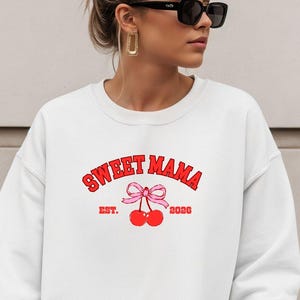 Może przedstawiać: Biała bluza z napisem "SWEET MAMA" w kolorze czerwonym, wygiętym nad różową kokardą i dwiema czerwonymi wiśniami. Pod wiśniami znajduje się napis "EST. 2020" w kolorze czerwonym. Bluza ma swobodny, wygodny styl.
