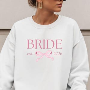 Op de afbeelding: Witte crewneck sweatshirt met het woord "BRIDE" in roze script, een roze strik afbeelding en het jaar "2026". De sweatshirt is een casual stijl, perfect voor een bruiloft.