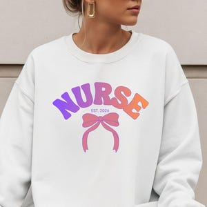 Peut inclure: Un sweat-shirt blanc avec le mot "NURSE" en dégradé violet à orange, orné d'un nœud rose. Le texte "EST. 2026" est au-dessus du nœud. Le sweat-shirt est de style décontracté.