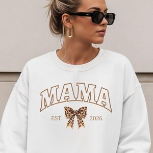 Może przedstawiać: Biała bluza z napisem "MAMA" w brązowych, łukowatych literach. Pod słowem "MAMA" znajduje się kokarda w panterkę i napis "EST. 2026". Bluza ma okrągły dekolt.