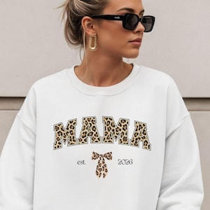 Może przedstawiać: Biała bluza z napisem "MAMA" w literach w panterkę. Poniżej kokarda w panterkę i napis "est. 2023". Bluza ma swobodny styl.