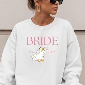 Peut inclure: Sweat-shirt blanc avec le mot "BRIDE" en rose, une colombe dessinée et l'année "2026". Le sweat-shirt est de style col rond. La colombe a des pieds jaunes et émet des ondes sonores.