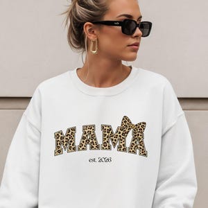Może przedstawiać: Biała bluza z napisem "MAMA" w panterkę na piersi. Pod napisem znajduje się tekst "est. 2023". Bluza ma długie rękawy i okrągły dekolt.