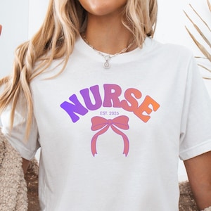 Peut inclure: T-shirt blanc avec le mot "NURSE" en violet et orange, un nœud rose et le texte "EST. 2026". Le t-shirt est porté avec un short en jean et un cardigan beige. Le modèle fait le signe de la paix.