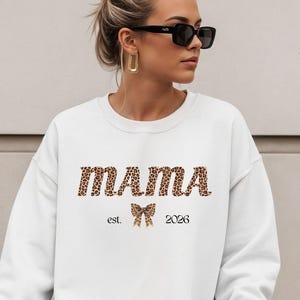 Może przedstawiać: Biała bluza z napisem "MAMA" w panterkę. Pod słowem "MAMA" znajduje się grafika kokardy i napis "est. 2026". Bluza ma długie rękawy i okrągły dekolt.