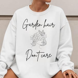 Puede incluir: Sudadera blanca con el texto "Garden hair Don't care" en fuente cursiva. La sudadera presenta un dibujo lineal de la cara de una mujer con una corona floral. La parte superior de manga larga es de estilo casual.