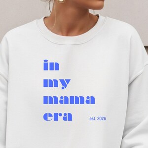 Puede incluir: Sudadera blanca con el texto azul "in my mama era est. 2026". El diseño es de estilo casual, adecuado para el uso diario. El texto está apilado verticalmente, con las palabras "in", "my", "mama" y "era" en negrita, sin serifa.