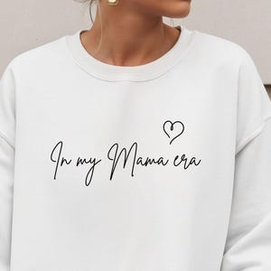 Może przedstawiać: Biała bluza z okrągłym dekoltem z napisem "In my Mama era" w ozdobnej czcionce, z małym symbolem serca. Bluza jest w jednolitym białym kolorze.