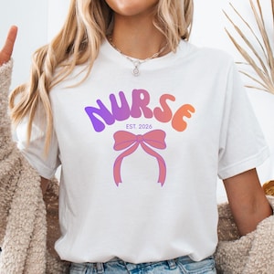 Pode incluir: Camiseta branca com a palavra "NURSE" em gradiente roxo e laranja, com um laço rosa. O texto "EST. 2026" está abaixo da palavra "NURSE". A camisa é usada com shorts jeans e um cardigan bege.