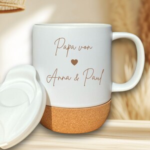 Pode incluir: Caneca de cerâmica branca com base de cortiça e tampa branca. A caneca tem uma alça e apresenta o texto "Papa von Anna & Paul" em escrita castanha, com um pequeno símbolo de coração. A tampa é colocada ao lado da caneca.