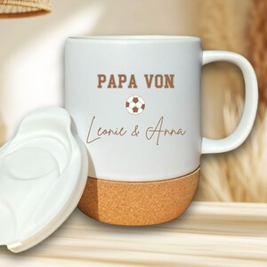 Pode incluir: Caneca de cerâmica branca com base de cortiça e tampa branca. A caneca apresenta o texto "PAPA VON" acima de um gráfico de bola de futebol, com os nomes "Leonie & Anna" abaixo. Ideal para bebidas quentes.