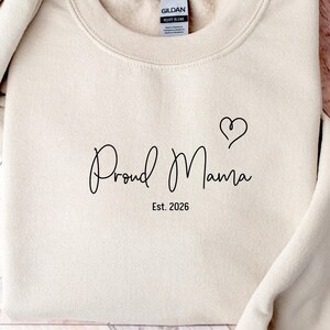 Może przedstawiać: Kremowa bluza z napisem "Proud Mama" w kursywie, symbolem serca i rokiem "Est. 2026" wydrukowanym na czarno. Bluza jest złożona, widoczne są rękawy.