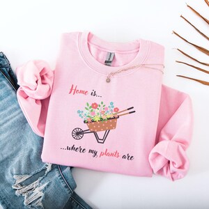 Puede incluir: Sudadera rosa claro con el texto "Home is... where my plants are". El diseño muestra una carretilla llena de flores coloridas. Un collar descansa sobre la sudadera, que se combina con unos vaqueros azules.