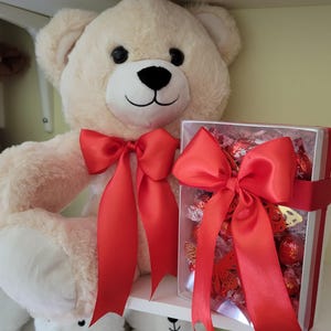 Puede incluir: Un oso de peluche color crema con un lazo de sat&eacute;n rojo y una caja de regalo llena de bombones. La caja de regalo est&aacute; atada con un gran lazo rojo. El oso tiene ojos negros y nariz negra.