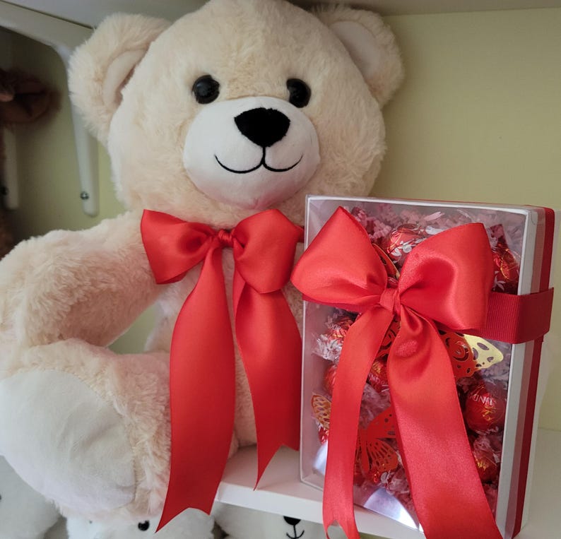 Puede incluir: Un oso de peluche color crema con un lazo de sat&eacute;n rojo y una caja de regalo llena de bombones. La caja de regalo est&aacute; atada con un gran lazo rojo. El oso tiene ojos negros y nariz negra.