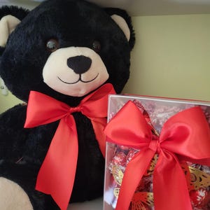 Puede incluir: Un gran oso de peluche negro con un lazo de sat&eacute;n rojo. Junto al oso hay una caja transparente llena de bombones y un lazo rojo. El oso tiene un hocico blanco y orejas beige.