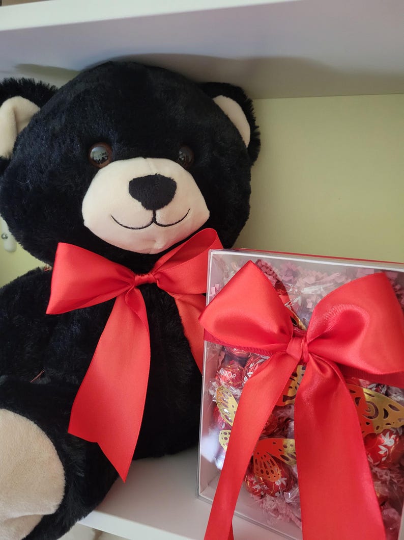 Puede incluir: Un gran oso de peluche negro con un lazo de sat&eacute;n rojo. Junto al oso hay una caja transparente llena de bombones y un lazo rojo. El oso tiene un hocico blanco y orejas beige.