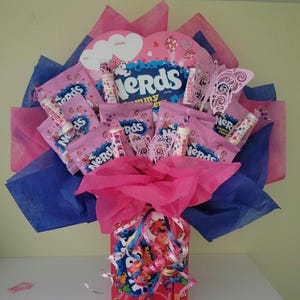 May include: A candy bouquet featuring pink Nerds candy packages, decorative butterfly cutouts, and heart-shaped accents. The arrangement is wrapped in pink and blue tissue paper, with a red box base.